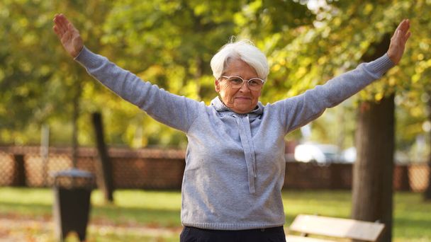 Senior Caucasian woman in glasses holds hands up doing sports gymnastics in the park healthy lifestyle concept middle shot selective focus park background  - Photo, Image