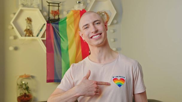 Glücklicher kaukasischer Schwuler, der in die Kamera lächelt, auf ein Regenbogenherz auf seinem T-Shirt zeigt, in seiner Wohnung posiert und vor der Regenbogenfahne steht. - Foto, Bild