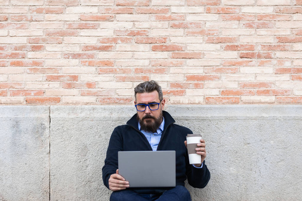 Yetişkin bir adam dışarıda duvara yaslanıp dizüstü bilgisayar kullanıyor ve elinde kahve fincanı tutuyor. - Fotoğraf, Görsel