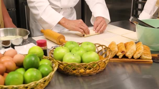 Großaufnahme an den Händen des Chefs in Kochuniform lehrt junge Kochkurs-Studenten Teig kneten und rollen, Zutaten für Backwaren zubereiten, Obstkuchen in der Edelstahlküche des Restaurants. - Filmmaterial, Video