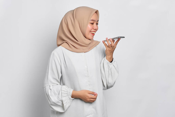 Улыбающаяся молодая азиатка-мусульманка с помощью голосового помощника на мобильном телефоне изолирована на белом фоне - Фото, изображение