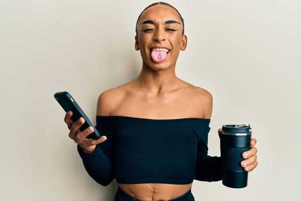 Hombre transgénero hispano usando maquillaje y cabello largo usando smartphone y bebiendo una taza de café sacando la lengua feliz con expresión divertida.  - Foto, imagen