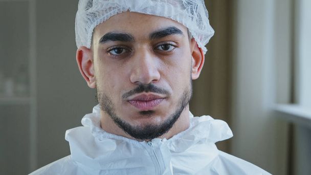Portrait of exhausted tired young arab male doctor nurse practitioner in protective uniform latex gloves stands in clinic takes off medical cap from head exhales with relief after hard day at work - Photo, Image