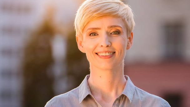 Portrait outdoors female happy smiling face natural makeup caucasian head with short hair mature 40s business woman standing posing outdoor in sunlight sunset sunbeams looking at camera with smile - Zdjęcie, obraz