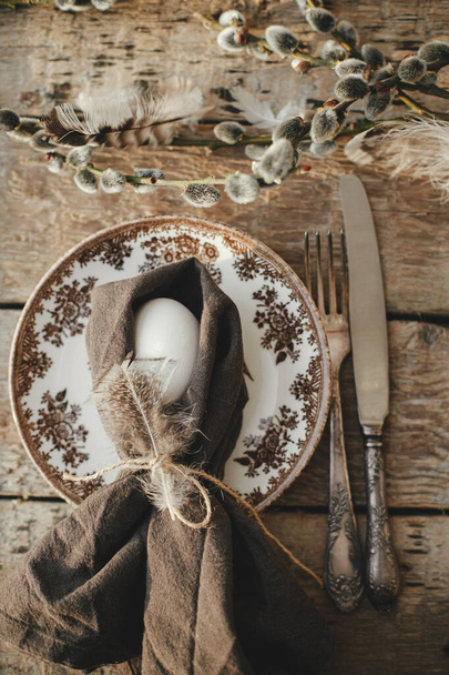Natural egg in napkin with feather, setting  with vintage plate, cutlery, pussy willow branches on aged wooden table. Rustic Easter table setting top view. Easter table decoration - Zdjęcie, obraz