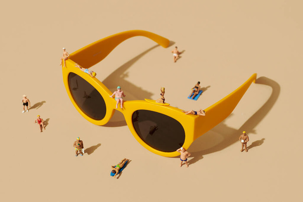 algunas personas en miniatura, con traje de baño, relajarse en la parte superior de un par de gafas de sol de plástico amarillo y algunas personas más en miniatura de pie alrededor, sobre un fondo marrón pálido - Foto, imagen