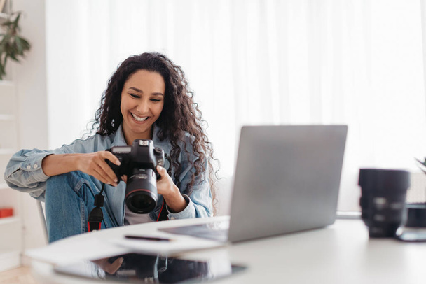 Χαρούμενος επαγγελματίας φωτογράφος Lady κρατώντας φωτογραφική μηχανή χρησιμοποιώντας το laptop στο χώρο εργασίας - Φωτογραφία, εικόνα