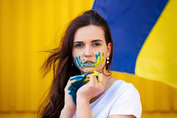 Mladá ukrajinská dívka pózuje s modrými a žlutými skvrnami na rukou a obličeji, ukrajinská vlajka na pozadí - Fotografie, Obrázek