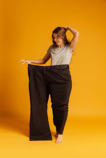 Portrait of young slim woman standing in giant black pants isolated over yellow background. Weight loss concept - Foto, Bild