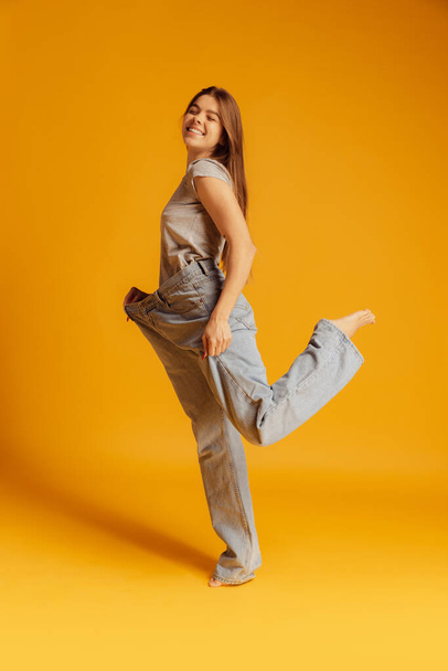 Portrait of young cheerful woman in big size jeans isolated over yellow background. Weight loss concept. - Φωτογραφία, εικόνα