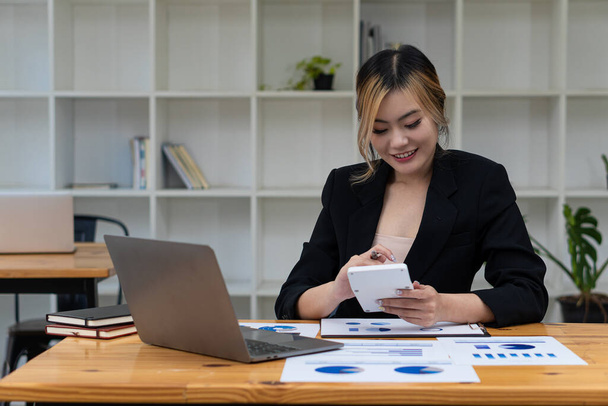 Gyönyörű ázsiai üzletasszony elemzése chart segítségével számológép, laptop az irodában papírok és a kezében toll, ül a széken a modern otthoni stúdióban. Mobil munkavállaló a vevőkkel - Fotó, kép