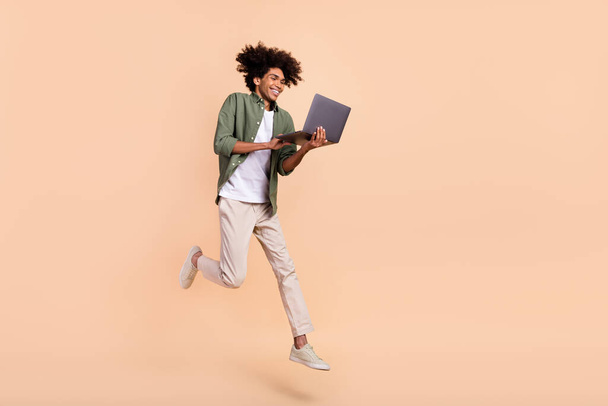 Full length body size view of attractive cheery trendy guy jumping using laptop work isolated on beige pastel color background - Foto, afbeelding