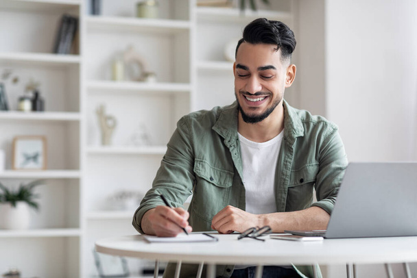 Sorrindo árabe masculino tomando notas enquanto trabalhava com laptop em casa escritório - Foto, Imagem