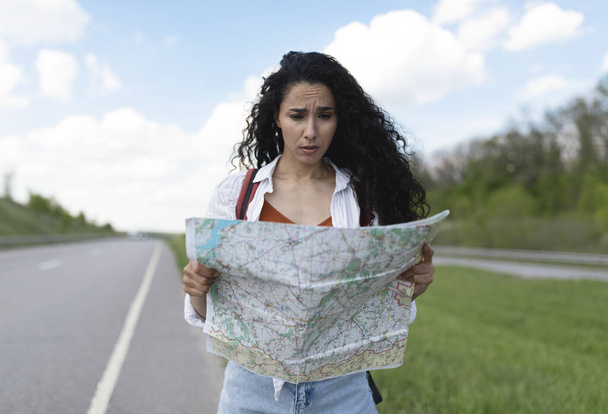 Zdezorientowana millenijna dama stojąca na drodze z mapą, zagubiona przy wyborze celu podróży, autostopem na przejażdżkę - Zdjęcie, obraz