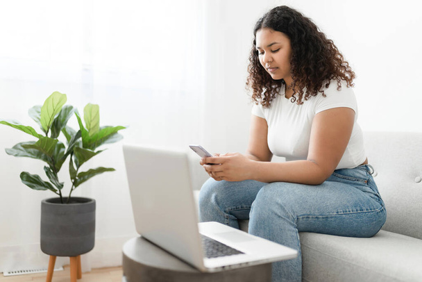 Attractive young African woman sitting on the couch at home with cellphone and laptop - Foto, afbeelding