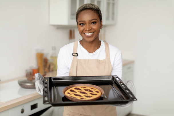 Froh lächelnde afrikanisch-amerikanische Frau in Schürze zeigt frischen leckeren Kuchen in minimalistischem Kücheninterieur - Foto, Bild