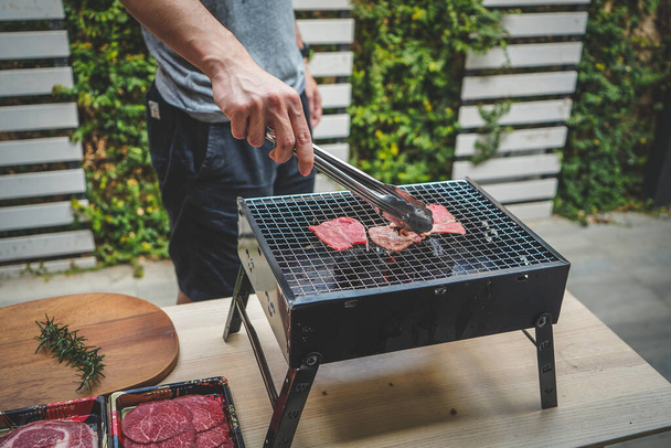Close up view of pieces of sliced beef on charcoal grill at Outdoor party. Human's hands using meat tongs to grill Multiple slices of premium meat on the stovetop. Home made barbecue beef grill smoke. - Foto, afbeelding