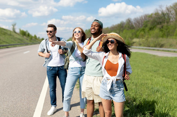 Ομάδα χαρούμενων φίλων που στέκονται στην άκρη του δρόμου, κάνοντας ωτοστόπ στις καλοκαιρινές διακοπές, πηγαίνοντας στην εξοχή, σταματώντας το αυτοκίνητο - Φωτογραφία, εικόνα