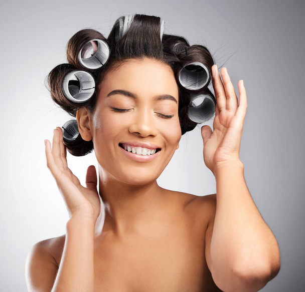 Putting her curls in. Studio shot of an attractive young woman posing with curlers in her hair against a grey background. - Foto, afbeelding