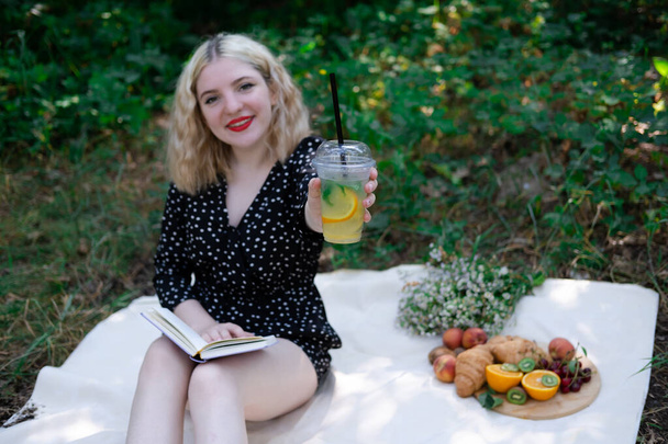 portrait of young woman on a picnic on plaid in park reading a book with tasty snacks. - Foto, imagen