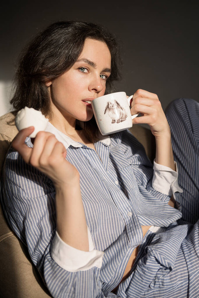 Portrait of a beautiful woman dressed in pajamas. She drinks coffee from a beautiful cup and holds a white gentle marshmallow. Filmed in the studio in the rays of morning sunshine. - Φωτογραφία, εικόνα