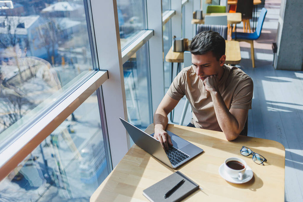 Genç ve çekici bir erkek serbest yazar bir kahve dükkanının içinde oturur ve pencereden dışarı bakar. Yakışıklı blogcu laptopta fikirler yazarken kafedeki tahta masada dizüstü bilgisayarla oturuyor. - Fotoğraf, Görsel