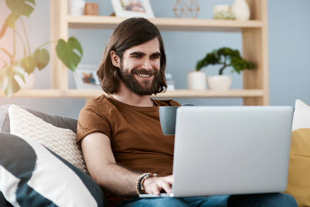 Jó kedve van ma. Egy jóképű fiatalemberről, aki a laptopját használja, miközben egy kanapén pihen a nappalijában.. - Fotó, kép