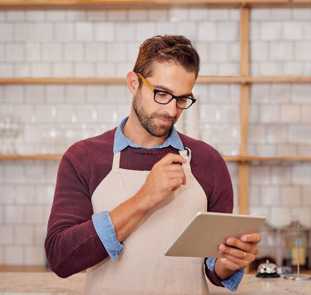 Hacer algunos cambios en el menú en línea. Shot de feliz joven empresario utilizando una tableta mientras está de pie en su cafetería. - Foto, imagen