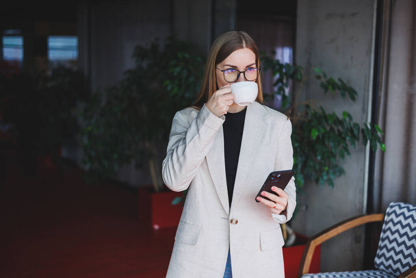 Pretty happy woman talking on a smartphone and drinking coffee while standing in a cafe. Young smiling blonde woman standing in a cafe. Concept of rest and free time. Modern female lifestyle - Foto, Imagen