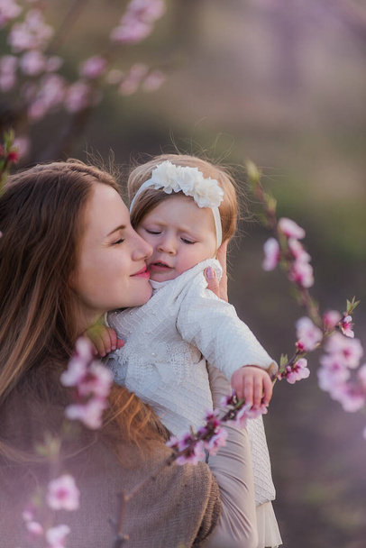 Young mother kissing little daughter in the blooming rose gardens. Portrait of woman hugging girl playing in nature among the trees. Single mother walks with her child in spring. Copy space - Valokuva, kuva