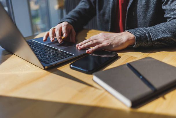 Ein Mann sitzt mit einem Laptop an einem Holztisch. Männliche Hände tippen in Großaufnahme auf einer Laptop-Tastatur. Konzept der jungen Menschen, die auf mobilen Geräten arbeiten. - Foto, Bild
