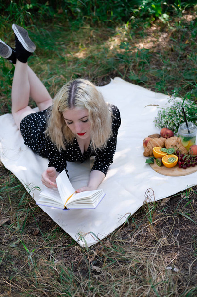 portrait of young woman on a picnic on plaid in park reading a book with tasty snacks. - Valokuva, kuva