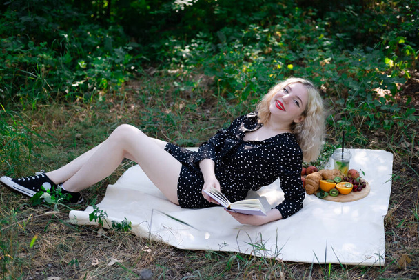 Porträt einer jungen Frau bei einem Picknick auf Plaid im Park, die ein Buch mit leckeren Snacks liest. - Foto, Bild