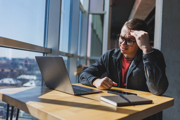 A freelancer in glasses looks into a laptop and he is angry while sitting at a table with a laptop and a notepad in a cafe during the day. Unlucky day. Working remotely from the office. - Photo, Image