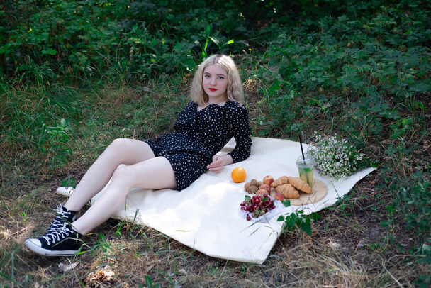 ładna blondynka na pikniku w lesie lub parku z lemoniadą, owocami i rogalikami. Lato, odpoczynek, relaks. pokolenie tysiącletnie. - Zdjęcie, obraz