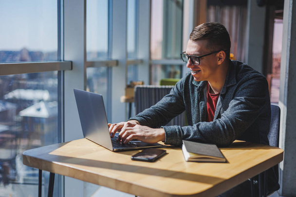 Portret van een man, IT-professional, werken op afstand met een moderne laptop, zitten aan een tafel en glimlachen naar de camera tijdens een pauze, een gelukkige menselijke programmeur in visie correctie bril - Foto, afbeelding