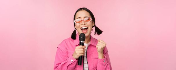 Щаслива красива азіатська дівчина співає з мікрофоном, насолоджуючись караоке, позує на рожевому студійному фоні
 - Фото, зображення
