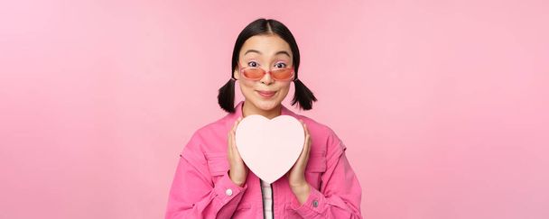 Kaunis aasialainen tyttö hymyilee onnellisena, näyttää sydämen lahjapakkauksen ja katselee innoissaan kameraa, seisoo vaaleanpunaisen romanttisen taustan yläpuolella - Valokuva, kuva