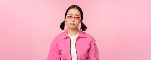 Πορτρέτο του λυπημένου και μελαγχολικού κοριτσιού από την Ασία μουτρώνει από την απογοήτευση, στέκεται αναστατωμένος σε ροζ φόντο στούντιο - Φωτογραφία, εικόνα