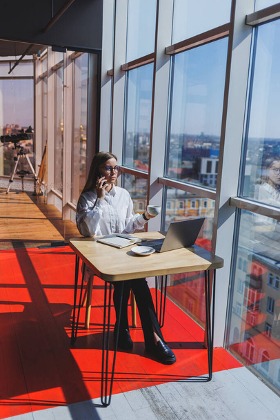 Empresaria gerente en una camisa blanca haciendo trabajo de oficina y sonriente, exitosa jefa europea en gafas ópticas para corrección de visión, posando en una mesa en un espacio de trabajo - Foto, imagen