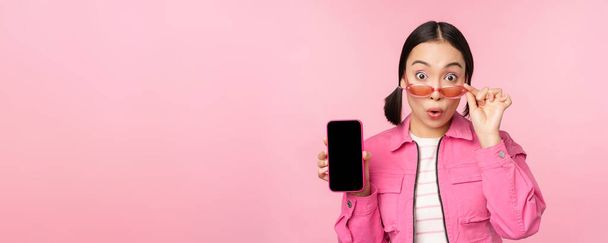 Portrait of asian girl showing mobile phone screen, reacting surprised, standing over pink background - Foto, imagen