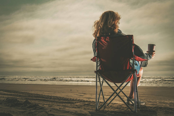Mujer caucásica con café en la mano disfrutando del tiempo libre en una playa. Sentarse en una silla de playa y ver atardecer. Tema Vida sin estrés. - Foto, Imagen