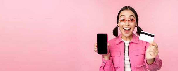 Asiatin zeigt Handybildschirm und Kreditkarte, reagiert überrascht auf Kamera, keucht beeindruckt, steht über rosa Hintergrund, Online-Shopping-Konzept - Foto, Bild