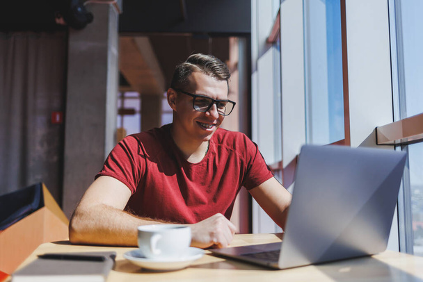 Portrait of a man, IT professional, working remotely with a modern laptop, sitting at a table and smiling at the camera during a break, a happy human programmer in vision correction glasses - Foto, afbeelding