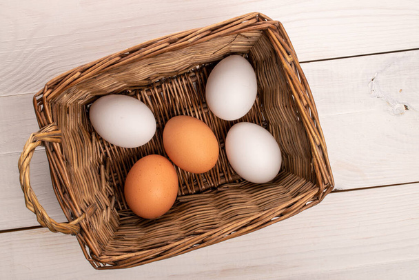 Varios huevos de gallina orgánicos frescos de color beige y blanco, en una canasta de mimbre, sobre una mesa de madera natural, pintados de blanco, vista superior. - Foto, imagen