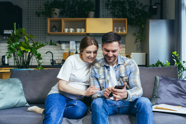 Young happy married couple husband and pregnant woman at home sitting on sofa using phone smiling - Foto, Bild