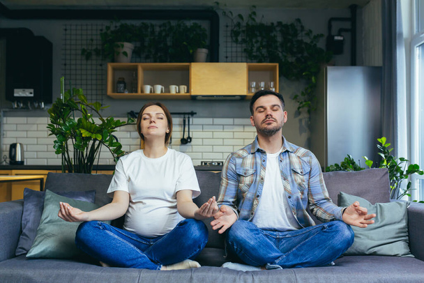 A pregnant man and woman are meditating in a lotus position on the couch - Foto, Imagem