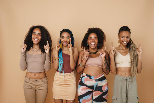 Young black four women laughing while pointing fingers upward together isolated over beige background - Foto, Bild