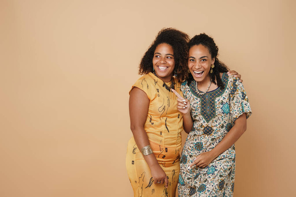 Jovem preto duas mulheres apontando o dedo para o lado e rindo da câmera isolada sobre fundo bege - Foto, Imagem
