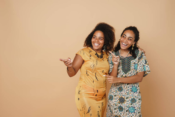 Young black two women pointing fingers aside and laughing at camera isolated over beige background - Foto, Imagen